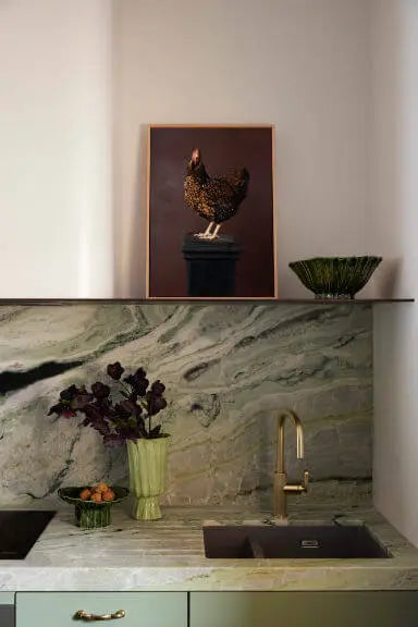 Fine art print of a brown speckled hen on dark background displayed above a marble kitchen counter with sage green cabinetry and brass fixtures