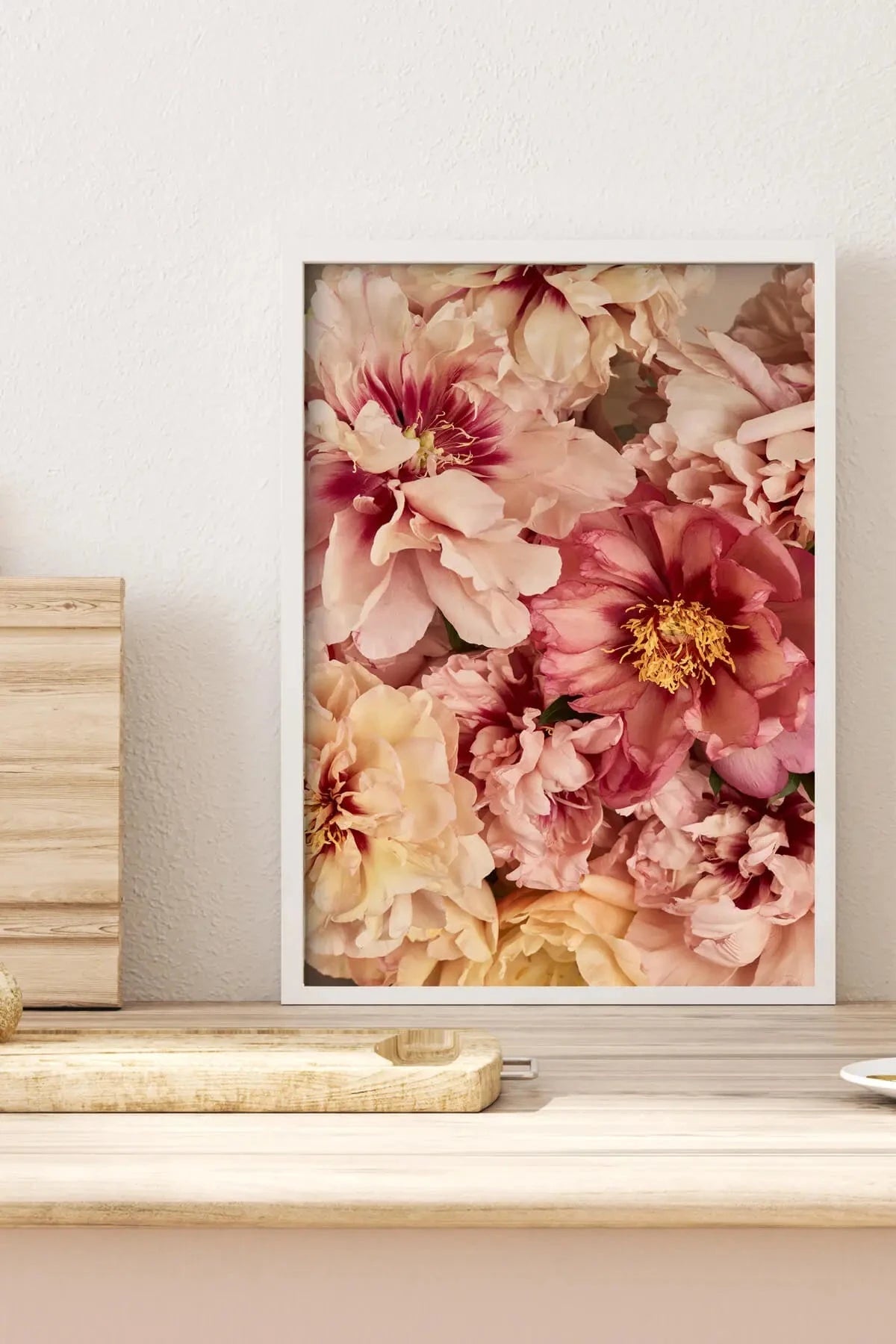 Fine art print of lush pink and cream peonies with golden centres, displayed in white frame on rustic timber shelf against neutral wall