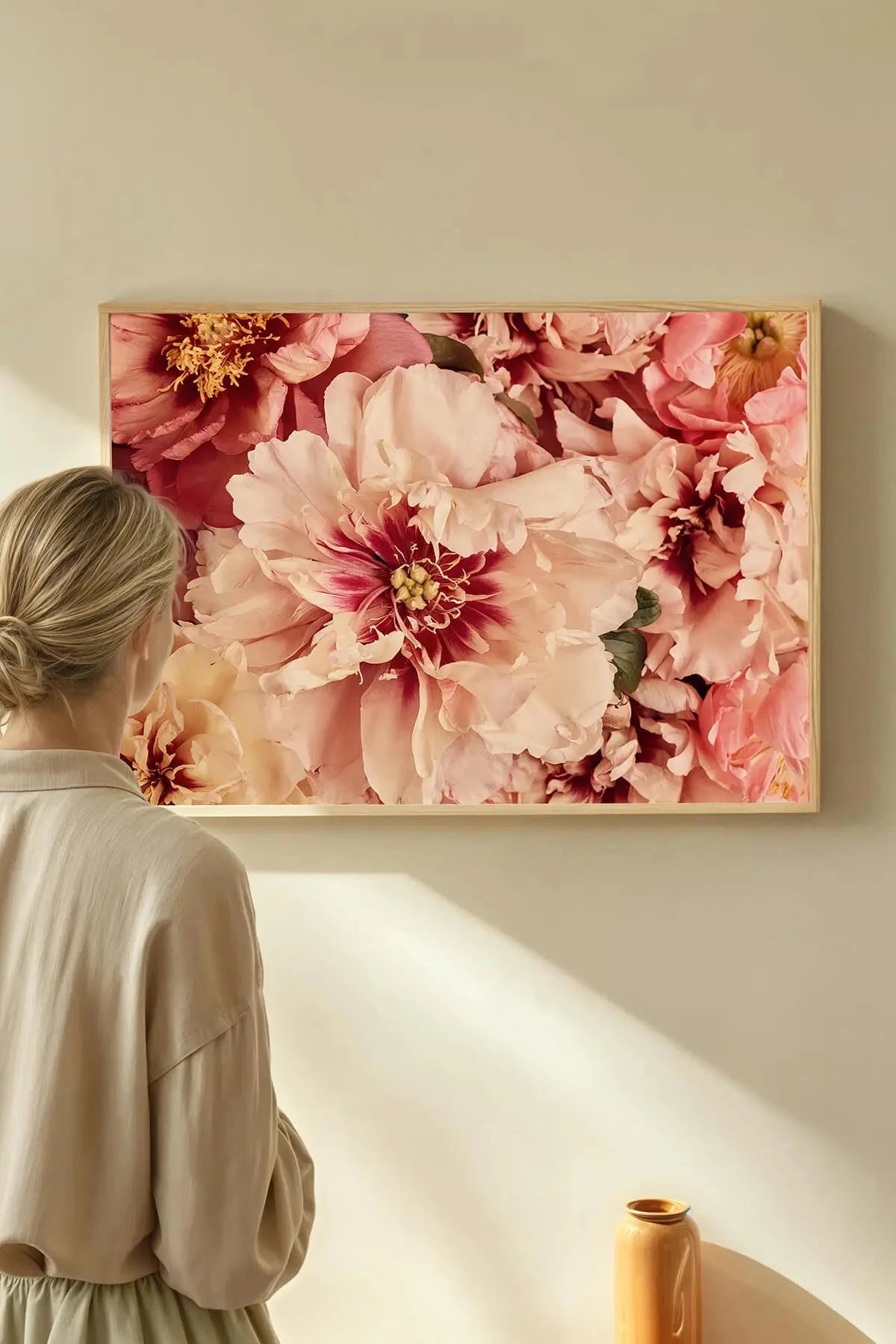 Fine art print of lush pink peonies with golden centers in a light timber frame, viewed by a person in a minimalist cream interior