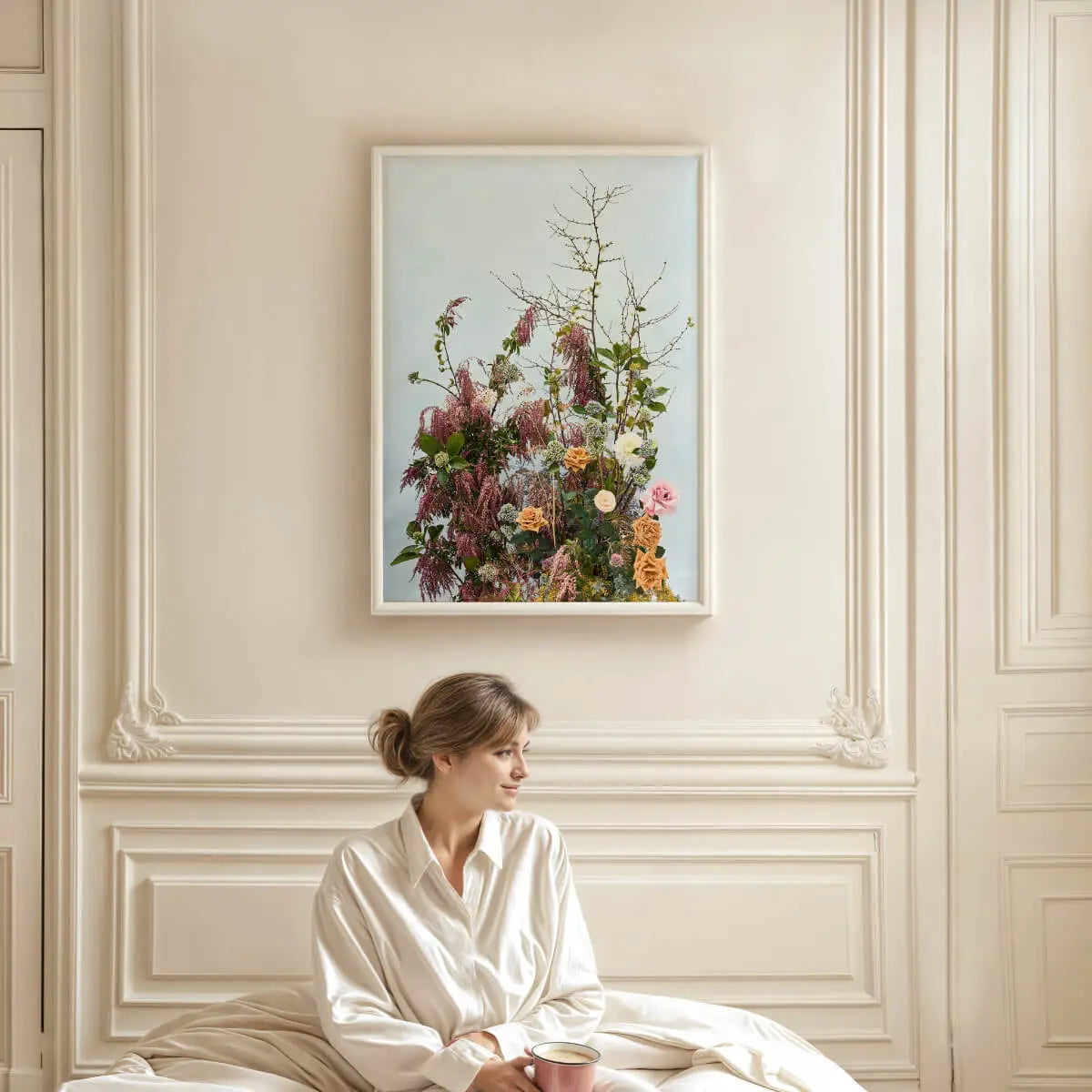 Botanical art print of wildflower arrangement with burgundy plumes and peach roses displayed above a woman in elegant French bedroom with cream paneled walls