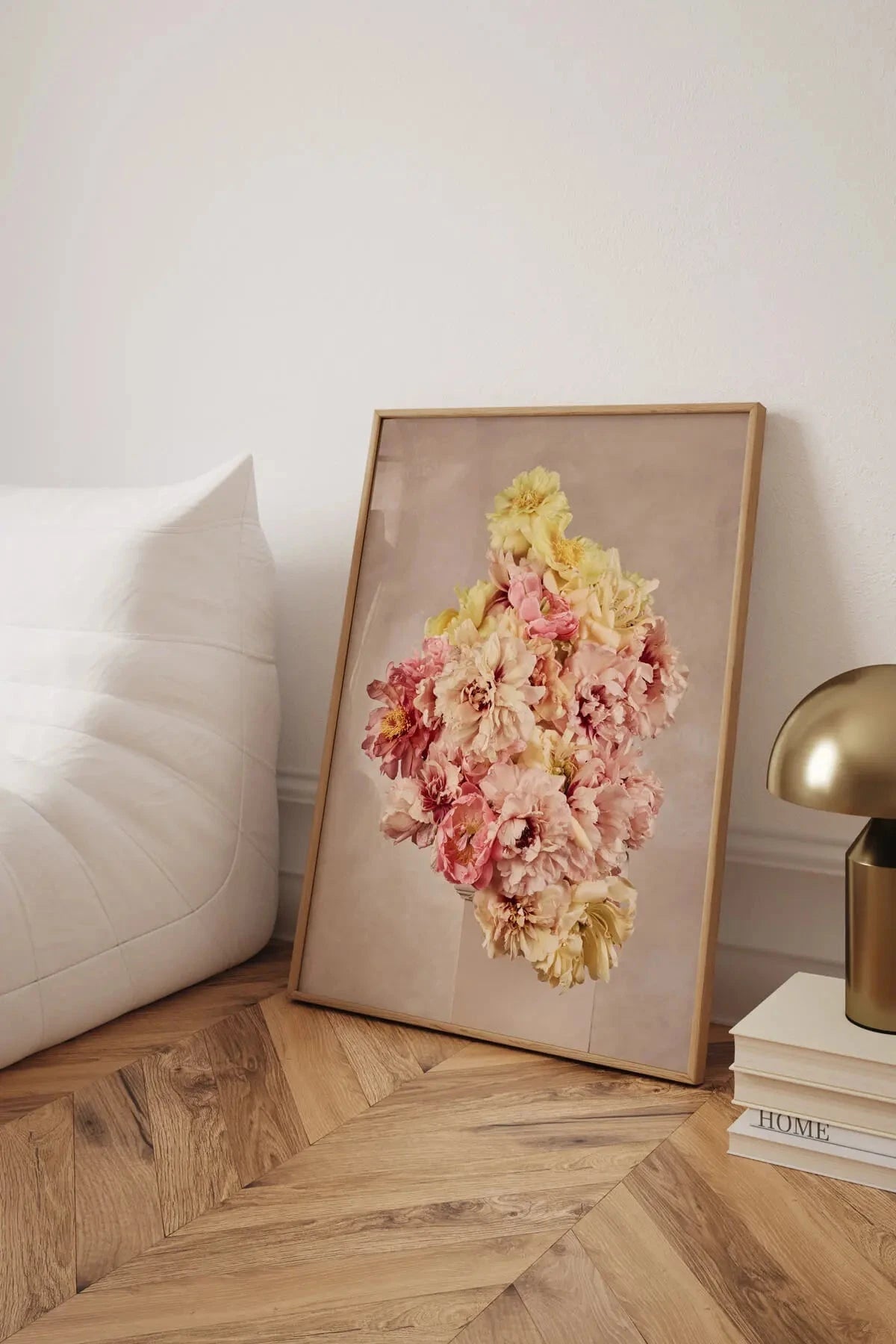 Botanical art print featuring pink and yellow peonies and carnations arranged in a vertical cluster, leaning against a white upholstered bed on herringbone timber flooring