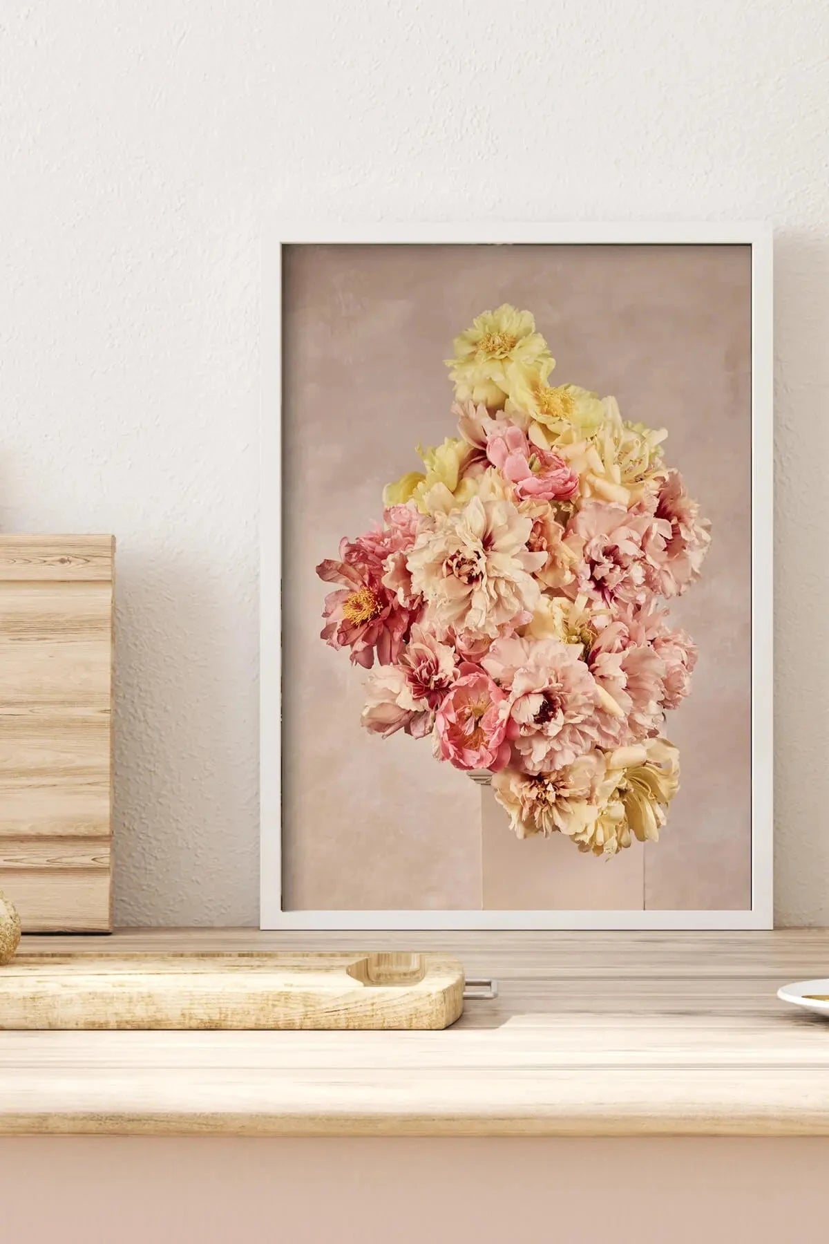 Fine art print of delicate pink and yellow peonies arranged vertically on a soft blush background, displayed on a minimalist shelf with natural timber accents.