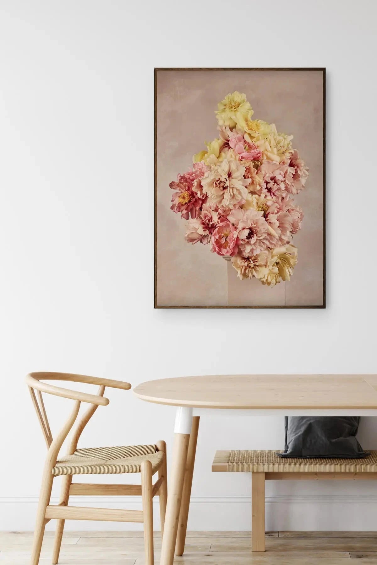 Fine art print of cascading pink, cream and yellow peonies on soft beige background, displayed above light timber dining table with woven chair
