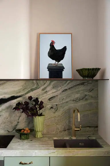 Fine art print of a black chicken with red comb perched on a plinth, displayed in a timber frame above a sage green kitchen with marble backsplash and brass fixtures.