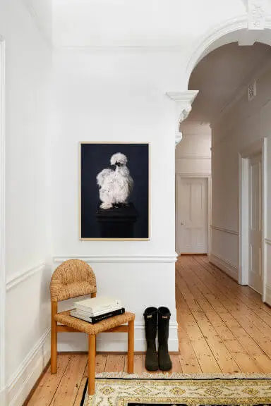 Fine art print of a white fluffy dog on a black background displayed in a bright hallway with white panelled walls, timber floors, and woven bench seat