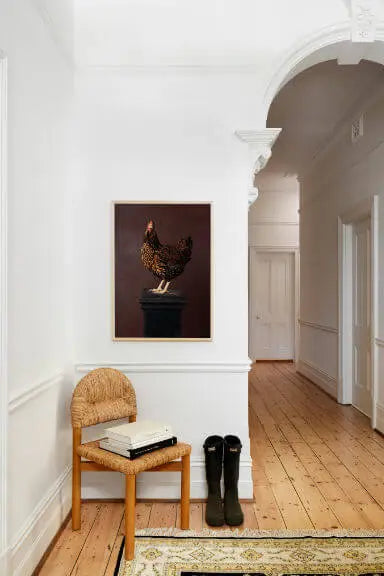 Fine art print of a brown speckled hen on dark burgundy background displayed above a woven chair in a bright hallway with white paneling and timber floors