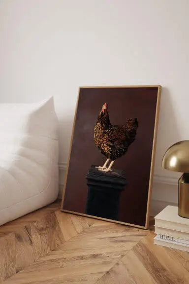 Fine art print of a speckled brown hen perched on a dark pedestal against a warm burgundy background, leaning against a cream sofa in a room with herringbone floors.
