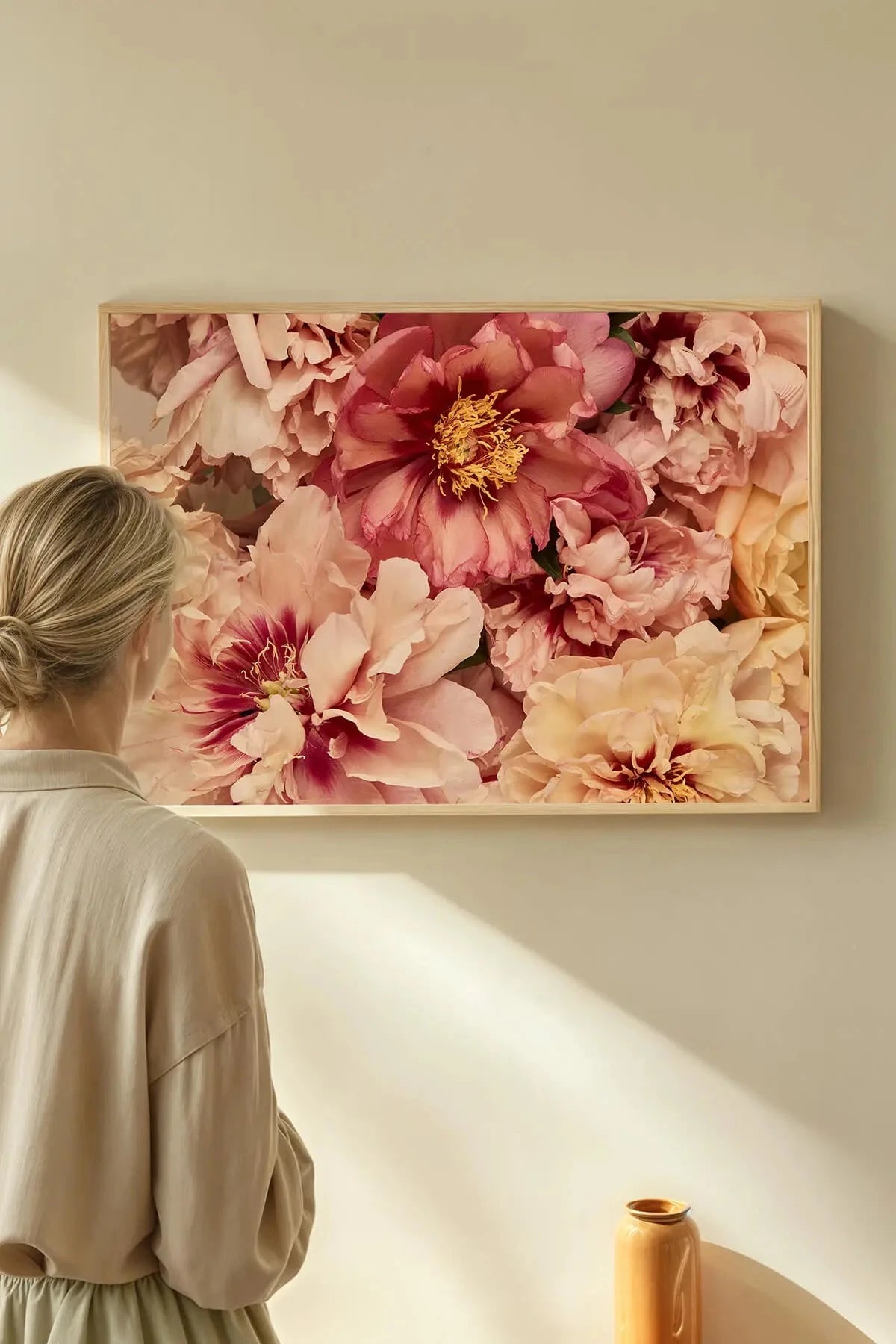 Fine art print of lush pink and coral peonies with golden stamens in a natural timber frame, displayed above a minimalist workspace with cream tones