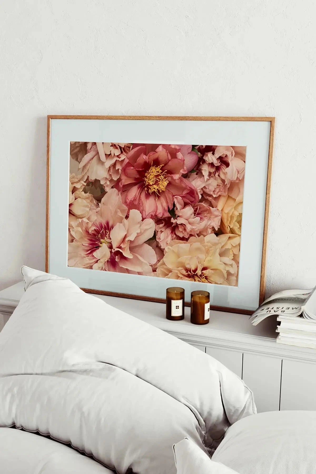 Fine art print of soft pink and coral peonies with golden centers in an oak frame, displayed above white bedding with amber candles on the nightstand.