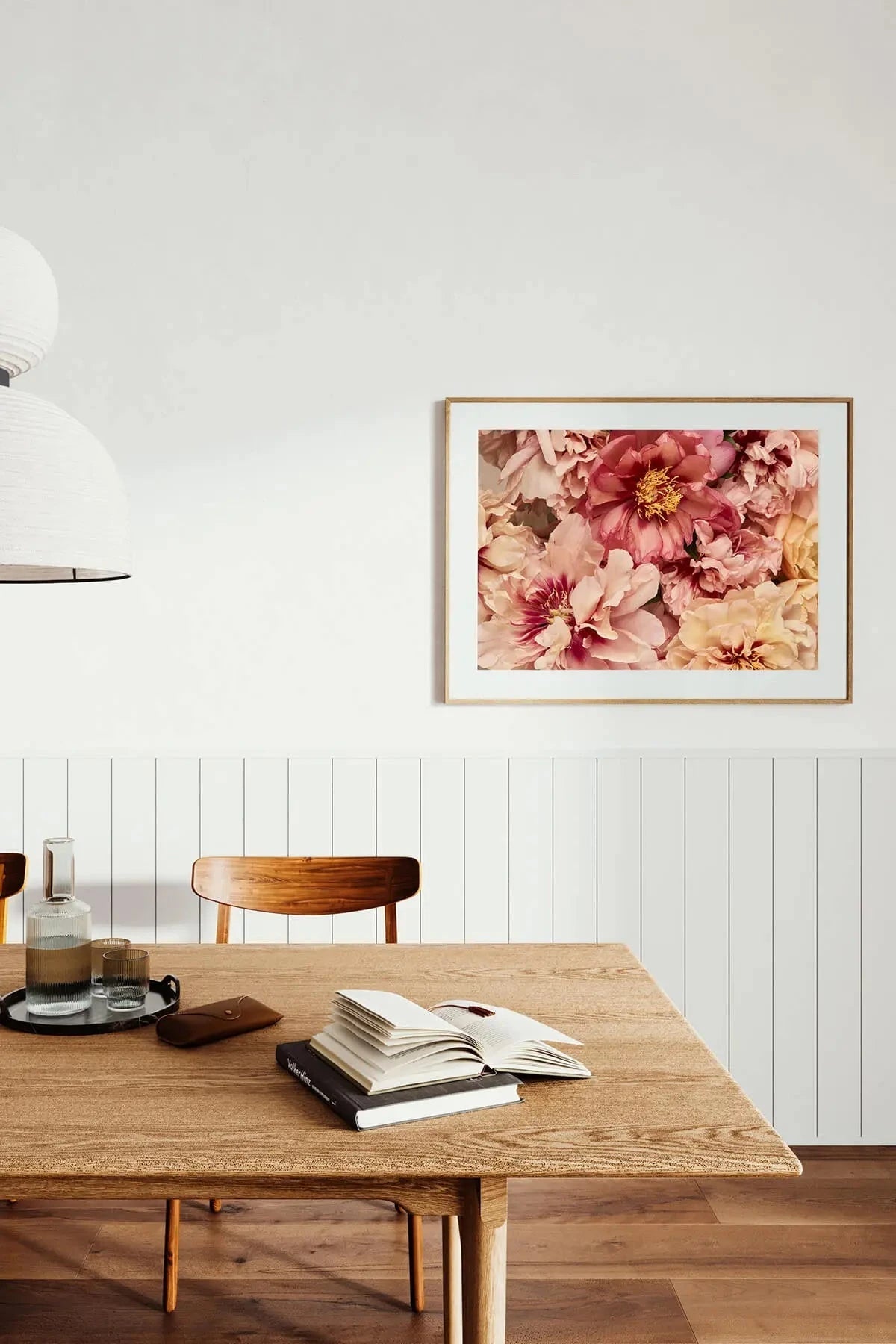 Fine art print of layered pink and coral peonies with soft petals and golden centers, displayed above a timber dining table in a minimalist white room.