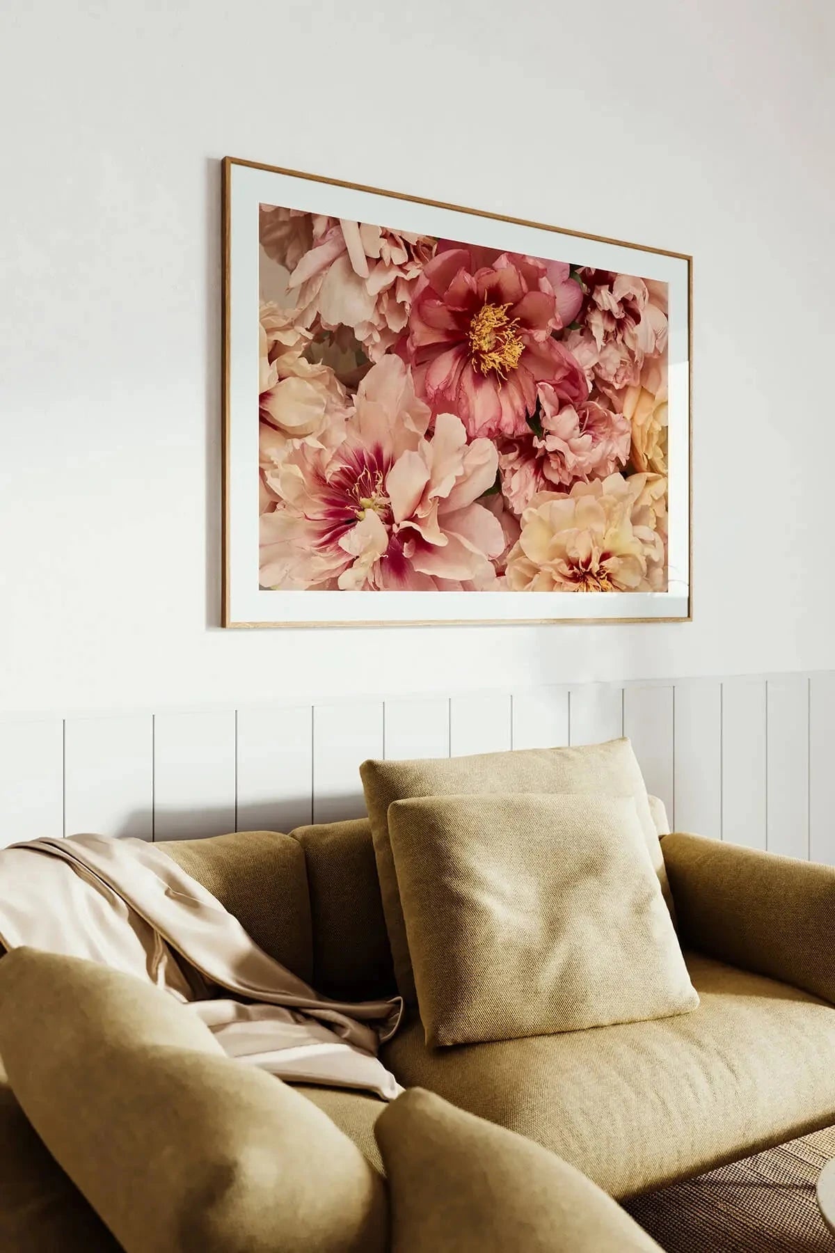 Fine art print of lush pink and peach peonies with golden centres in a timber frame above an olive green sofa against white panelling.