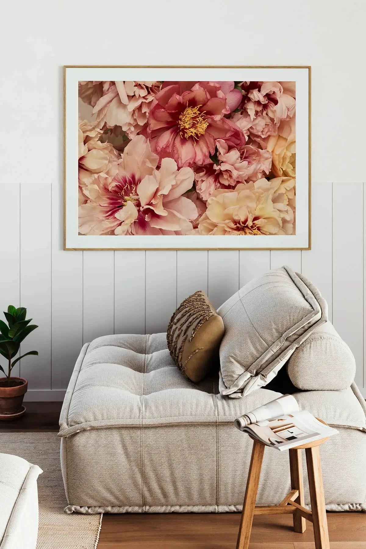 Fine art print of lush pink and cream peonies with golden centres in a natural timber frame, displayed above a neutral linen sofa in a coastal-style living room.