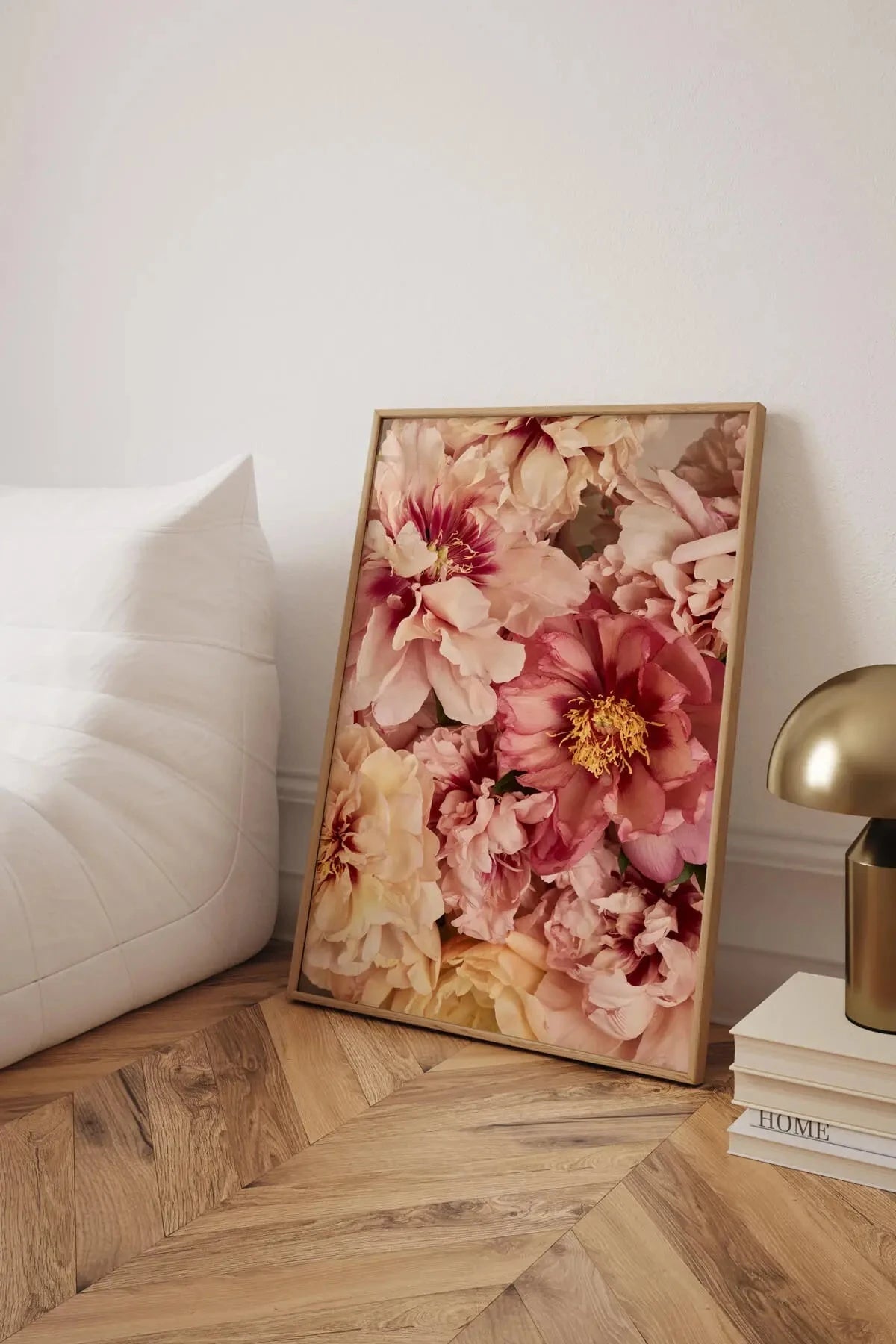 Fine art print of lush pink and cream peonies with golden stamens leaning against a white upholstered bed in a bedroom with herringbone timber floors and brass lamp