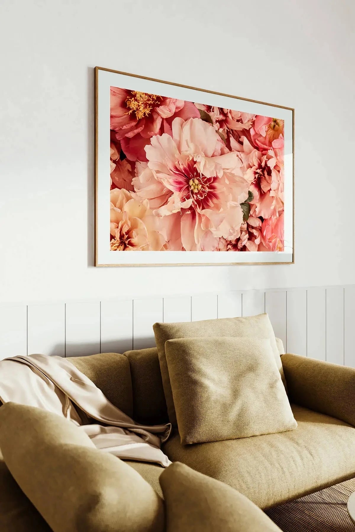 Fine art print of lush coral and blush pink peonies in a gold frame, hanging above an olive green velvet sofa against white panelled walls.