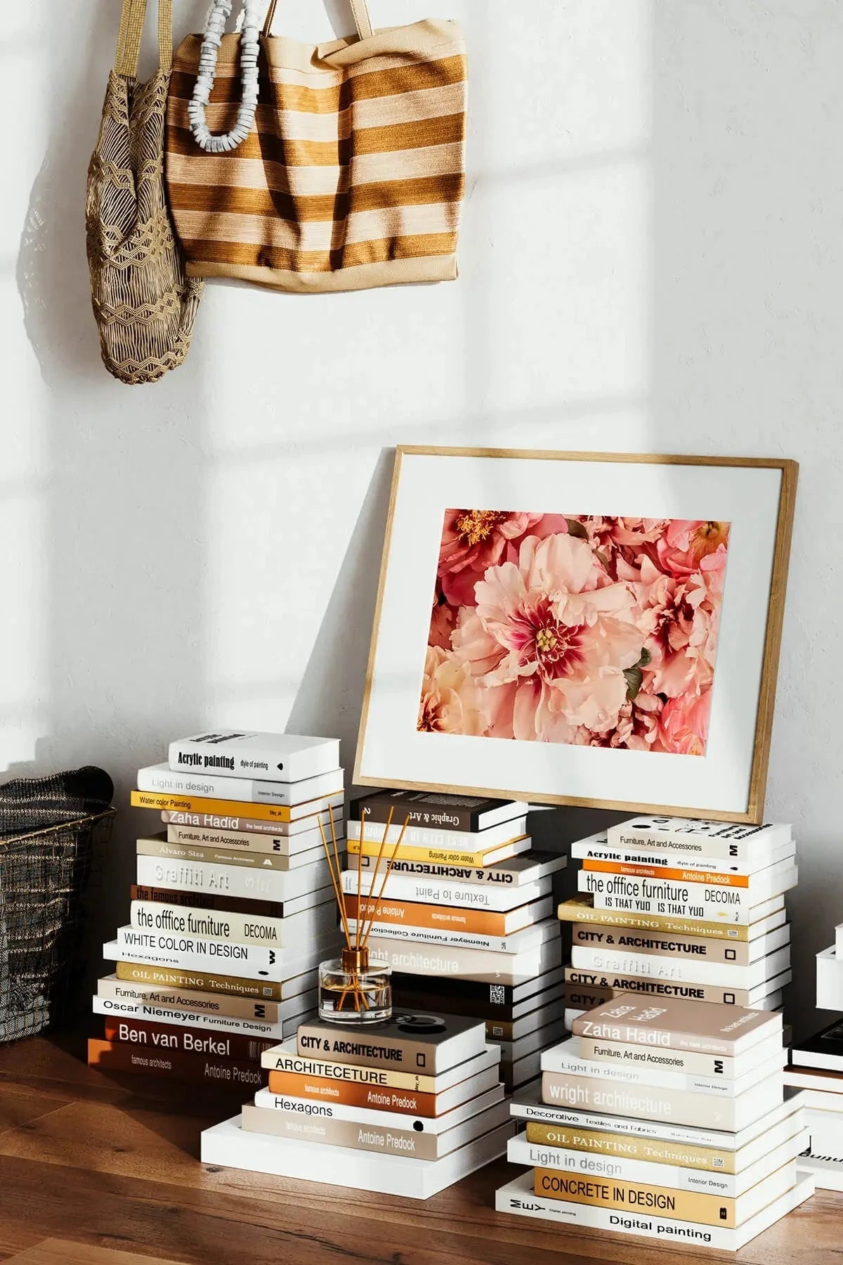 Fine art print of pink and coral peonies in full bloom, displayed in a timber frame on stacked design books beside a reed diffuser in a sunlit interior