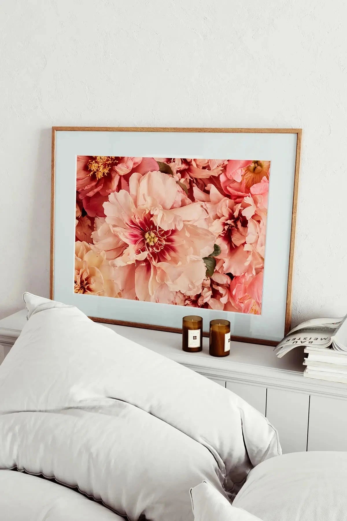 Fine art print of lush pink and coral peonies with golden stamens in an oak frame, styled above white bedding with amber candles on a bedside table.