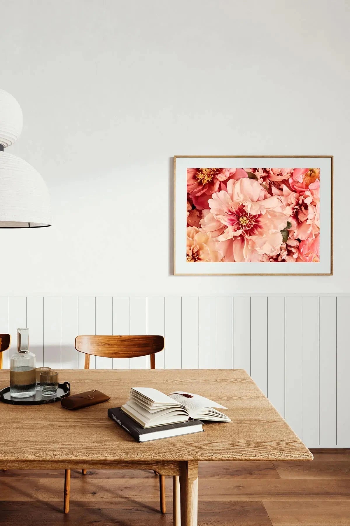 Botanical art print of lush pink and coral peonies in full bloom displayed above a wooden dining table in a minimalist room with white panelling.