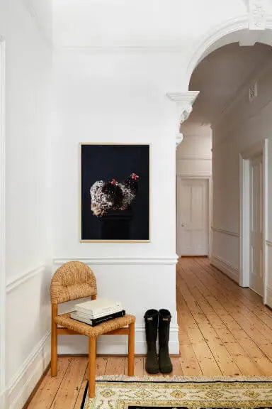 Fine art print of a floral wreath with pink roses on a black background, displayed in a bright hallway with white walls, wooden floors, and a woven chair beneath.